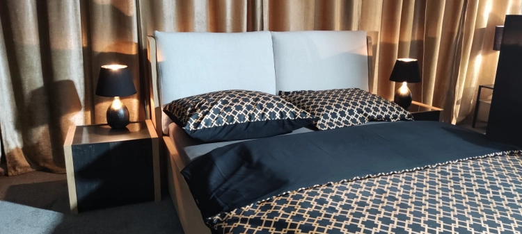 Łóżko MODELLO z tapicerowanym zagłówkiem New Elegance. Łóżko z miękkimi poduchami na odchylonym wezgłowiu, w tkaninie Positano o strukturze delikatnej plecionki z kolekcji Davisa.