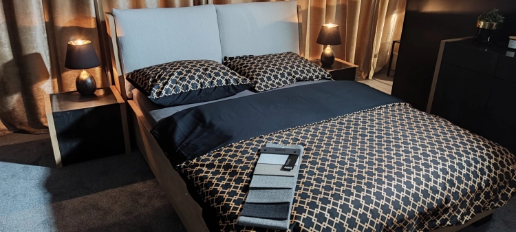 Łóżko MODELLO z tapicerowanym zagłówkiem New Elegance. Łóżko z miękkimi poduchami na odchylonym wezgłowiu, w tkaninie Positano o strukturze delikatnej plecionki z kolekcji Davisa.