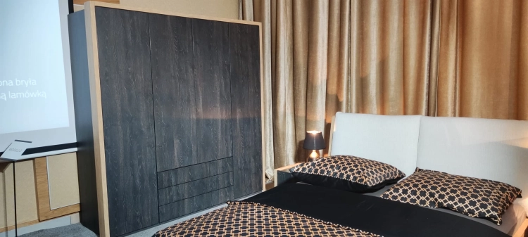 Łóżko MODELLO z tapicerowanym zagłówkiem New Elegance. Łóżko z miękkimi poduchami na odchylonym wezgłowiu, w tkaninie Positano o strukturze delikatnej plecionki z kolekcji Davisa.